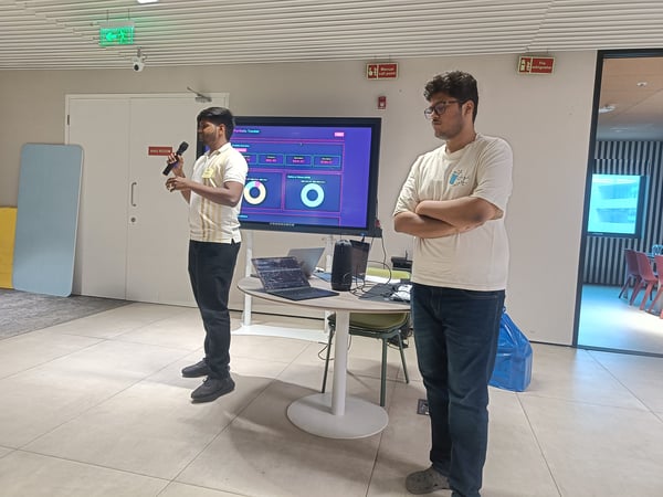 Two young men are giving a presentation in an office setting, with one speaking into a microphone while standing next to a large monitor displaying a data dashboard.