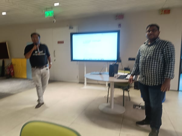 A photograph showing two men in an indoor setting, possibly during a presentation or demo. One man is holding a microphone while standing near a large screen, and another man stands closer to the camera.