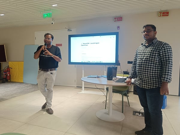 Two men are conducting a presentation in a modern indoor space, with one man speaking into a microphone next to a large display screen showing software details.