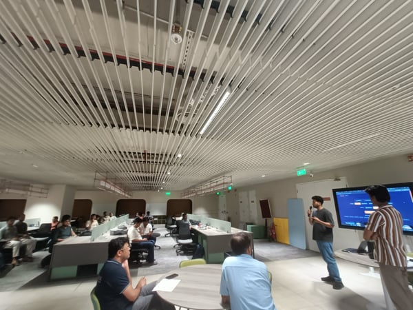A man is giving a presentation in a modern office setting, using a large digital screen while a group of people listens from their desks.