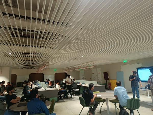 A group of people are gathered in a modern office space for a presentation or demo, with one person standing and speaking near a large screen.