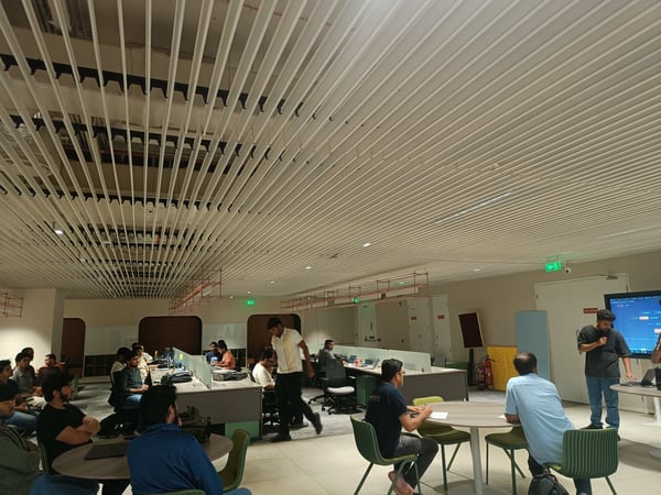 A group of people are gathered in a modern, open-plan office space for a presentation or demonstration. One individual stands near a large screen while others sit at desks and tables, observing.