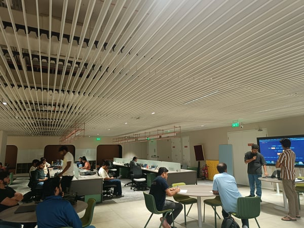 A group of people are gathered in a modern office or coworking space with a distinctive slatted ceiling, some working at desks and others attending a presentation near a large screen.