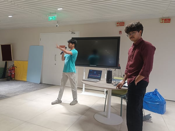 Two young men are in a modern indoor setting; one is speaking into a microphone during a presentation, while the other stands beside a table with a laptop and a large monitor.
