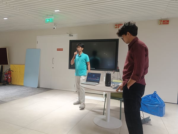 Two young men are in a modern indoor space where one is speaking into a microphone during a presentation, while the other stands nearby observing.