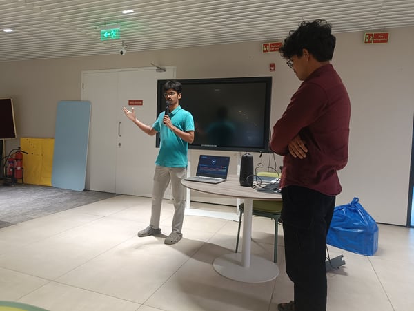A young man in a teal polo shirt is giving a presentation using a microphone and a large screen, while another man in a maroon shirt observes from the side.