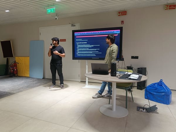 Two men are in an indoor setting where one is giving a presentation using a microphone and a large screen displaying a SWOT analysis.