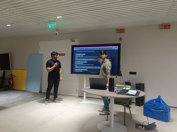 Two young men are conducting a presentation in a modern indoor setting. One man holds a microphone while the other points to a large screen displaying text about different business perspectives.