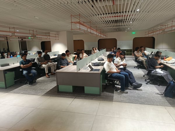 A group of people are seated at desks in a modern, well-lit office space, seemingly attending a presentation or meeting.