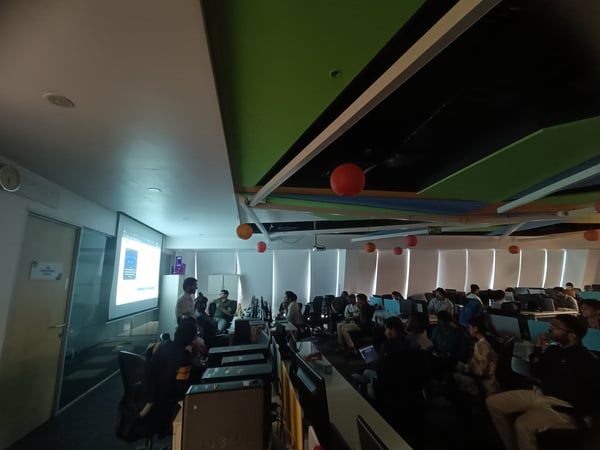 A group of people in a modern office or coworking space, with one person giving a presentation on a projector screen while others sit at desks with computers.