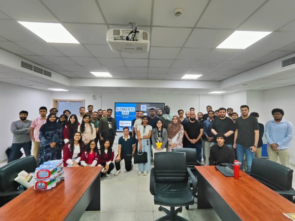 A group photograph of attendees posing together in a classroom or seminar room, with a screen in the background displaying 'AI Tinkerers Dubai'.