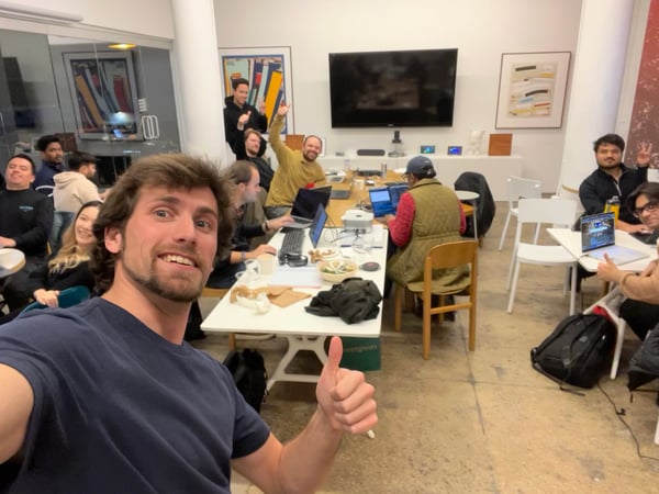 A group of people are gathered in a modern office or coworking space, with a man in the foreground taking a selfie and giving a thumbs-up. Several individuals are sitting at long white tables with open laptops, appearing to be working or collaborating in a casual environment.