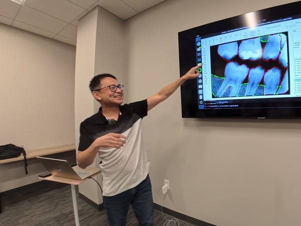 A man in a black and white polo shirt and glasses stands in an office setting, smiling and pointing at a large monitor displaying a dental X-ray annotated with colorful segmentation lines.