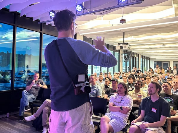 A man wearing a camera strap addresses a large, seated audience in a modern indoor venue, characterized by large windows overlooking a waterfront scene at twilight.