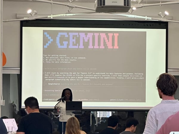 A photograph capturing an indoor technical presentation where a speaker, a woman, stands before a large screen displaying a command-line interface titled "GEMINI" and related technical text, while an audience watches.