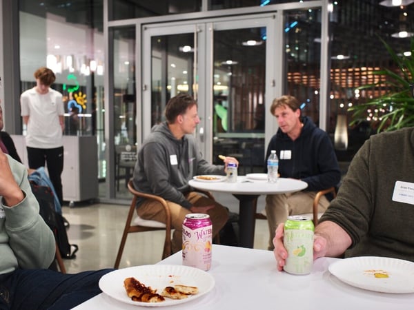 A candid photo captures a group of men socializing and eating snacks at white tables during an informal indoor evening event, likely a networking session in a modern office space.