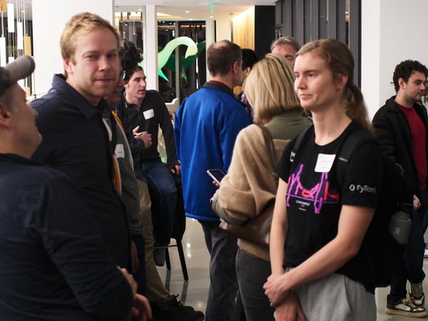 A candid photograph captures a group of people socializing and networking indoors at what appears to be a professional or tech conference event, indicated by name tags and a 'PyTorch' branded shirt.