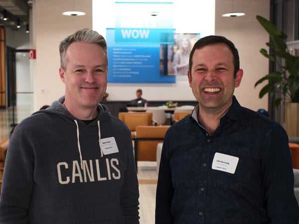 A photograph captures two men smiling directly at the camera in a brightly lit, modern indoor lounge or event space, both wearing name tags.
