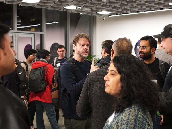 A candid photograph captures a group of people networking and conversing indoors at an event, featuring a man in the center speaking and gesturing while others listen or interact nearby.