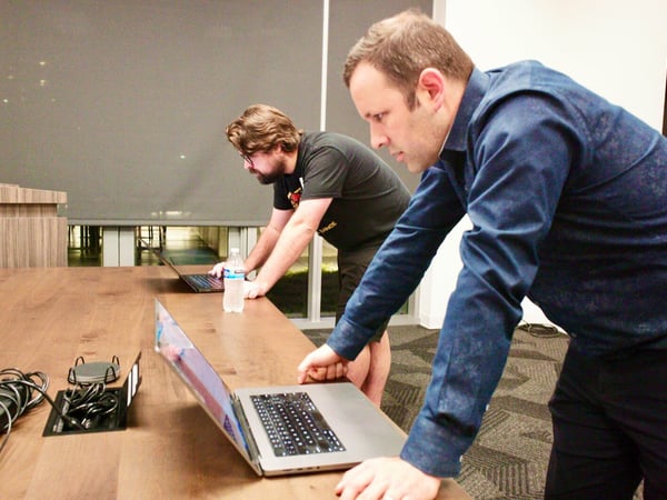 Two men lean over a long wooden table in a conference room, intently focused on their open laptops, possibly preparing for a presentation or debugging equipment.