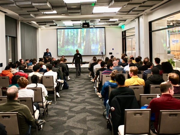 A wide-angle photograph captures a large audience seated in rows, attending a presentation or talk in a modern conference room. A large screen at the front displays an image of a forest, and two men are visible near the stage area.
