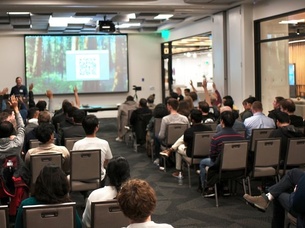 A wide-angle photograph captures a large audience seated in a conference room attending a presentation, with many attendees raising their hands, likely participating in a Q&A session. A projection screen at the front displays a QR code over a background image of a forest.