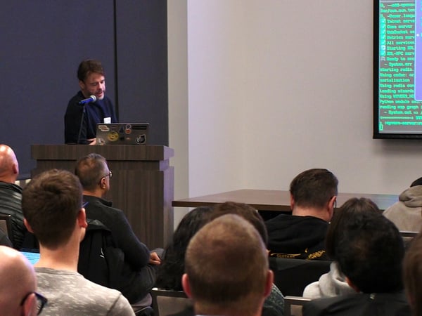 A photograph capturing a technical presentation where a man is speaking at a podium while an audience sits facing him. A large screen displays lines of code and terminal output in green text on a dark background.