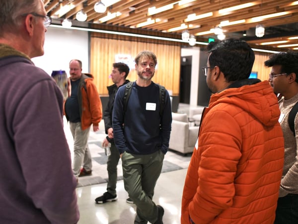 A candid photograph captures a group of six men networking or socializing in a modern indoor venue, characterized by wood-slat ceilings and warm lighting.