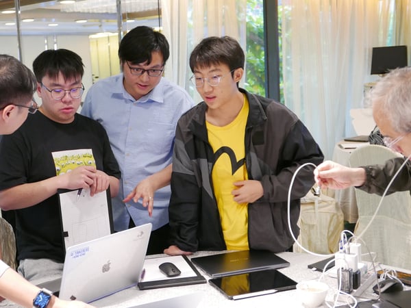 A group of young men are gathered around a table with laptops and papers, engaged in a collaborative discussion or project work.