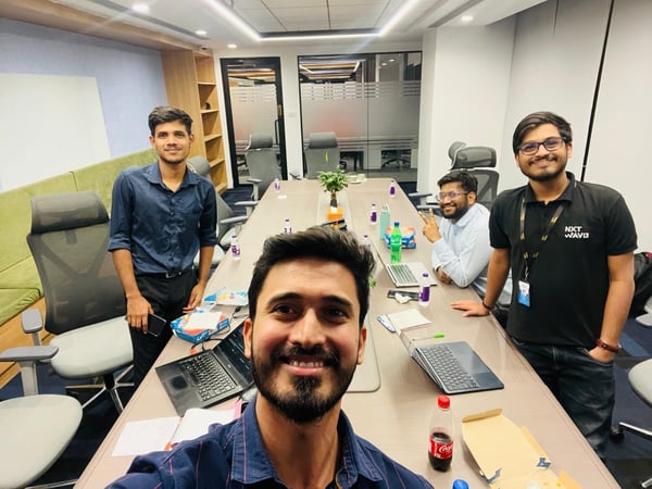 A group selfie captures four young men gathered around a long, polished conference table in a modern office meeting room, surrounded by laptops, drinks, and snacks.