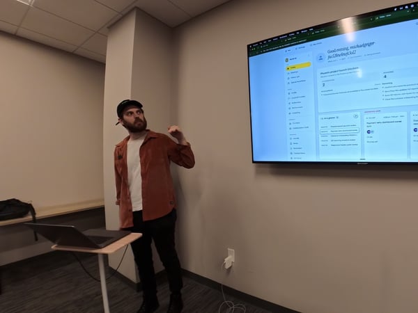 A man in a brown button-up shirt and a black cap stands in an office setting, pointing toward a large wall-mounted TV screen displaying a digital project management dashboard.