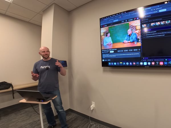 A man wearing a grey NASA t-shirt stands next to a laptop stand, presenting in front of a large wall-mounted TV screen displaying a web application interface with video and transcript elements.
