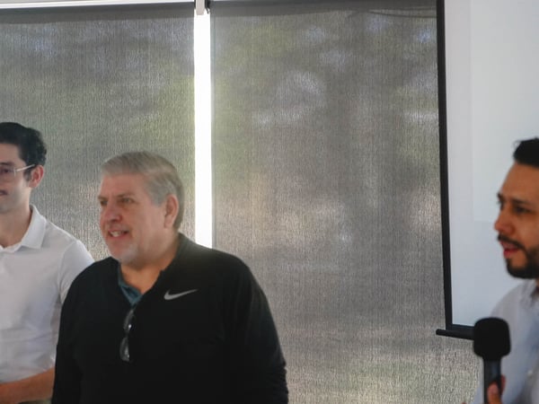 The image shows three men at an indoor event, with one man on the right speaking into a microphone while the others stand behind him.