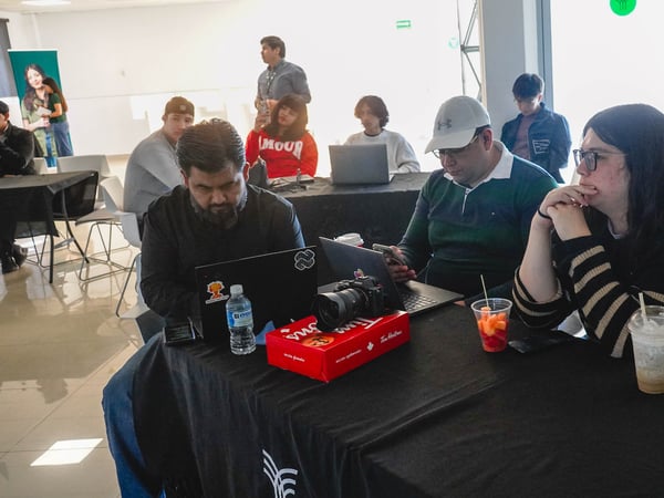 A group of people are gathered around a table in a bright indoor space, some working on laptops and others observing. The scene includes snacks like Tim Hortons donuts and drinks, suggesting a casual workshop or event.