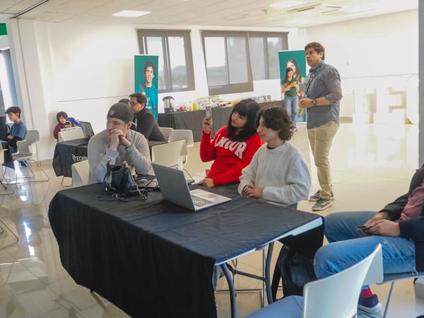 A group of young people are gathered in a bright indoor space, likely for a tech-related event or workshop, with some sitting at tables with laptops and others standing in the background.