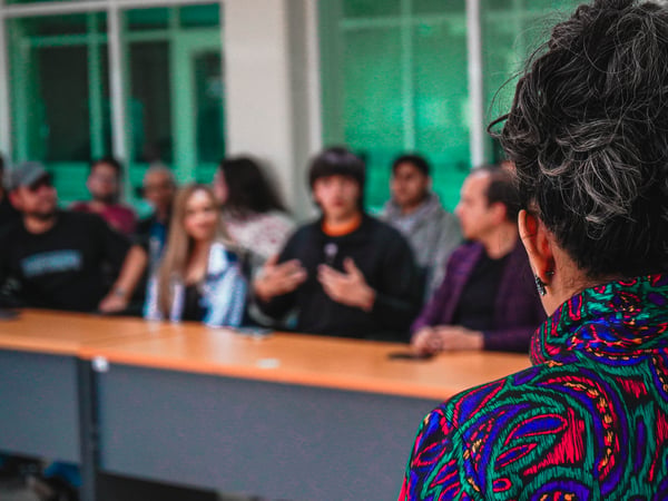 A woman with her hair in a bun and wearing a vibrantly patterned shirt is seen from behind, facing a group of people seated at long tables for a discussion or presentation.