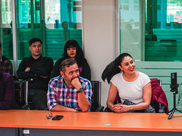 A group of people are seated at a long orange table in an office environment, appearing to be attending a presentation or meeting.