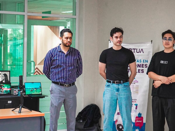 Three young men stand in a room, appearing to be part of a presentation or demo event. To their left is a desk with computer monitors and a tripod, while a banner and projection screen are behind them.