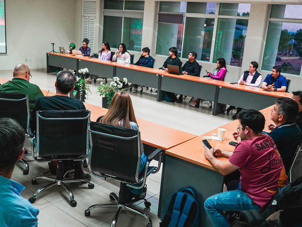 A group of people are gathered in a modern meeting room for a presentation, seated around a U-shaped table arrangement while looking towards a projector screen.