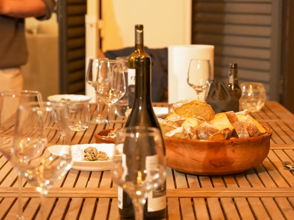 A wooden table is set for a social gathering, featuring wine bottles, several glasses, and a large ceramic bowl filled with sliced bread.