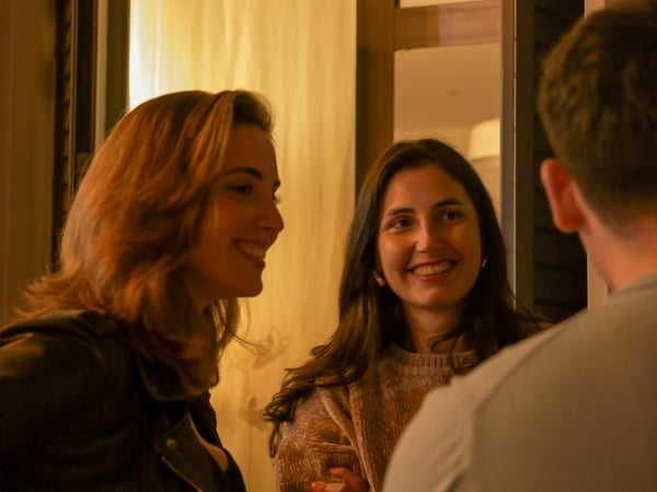 A candid indoor photograph showing two women smiling and engaging in a conversation with a man whose back is to the camera.