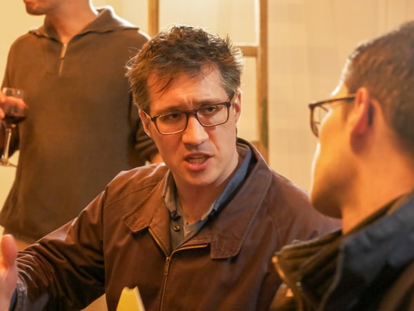 A man with glasses is captured in a candid moment, engaged in an earnest conversation with another person in a warm, indoor setting.