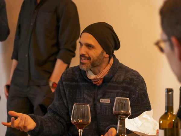 A man wearing a beanie and a fleece jacket is captured mid-conversation, gesturing with his hand while sitting at a table with wine glasses.