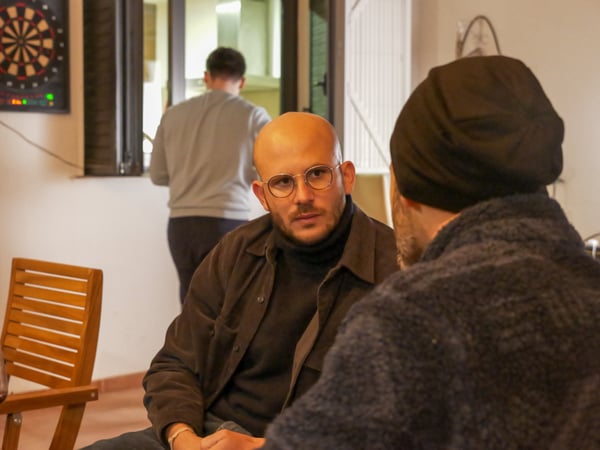 A man with glasses and a beard is engaged in a conversation with another person whose back is to the camera, while a third person stands in the background near a window.