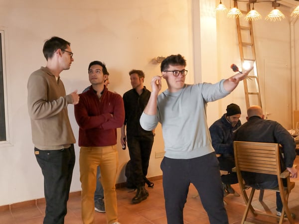 A group of men are gathered in an indoor setting with warm lighting, where one man in the foreground is captured in the middle of throwing a dart.