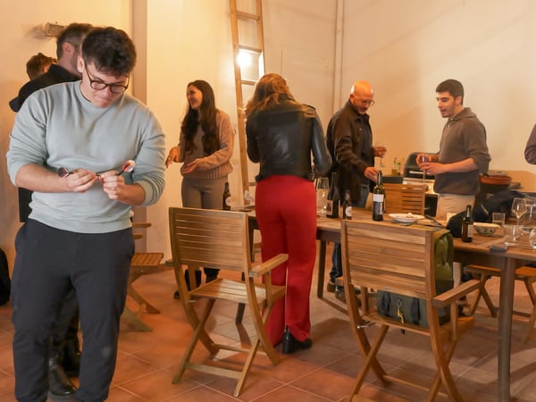 A group of people are gathered in an indoor social setting, with some standing around a wooden table with drinks and others interacting in the background.