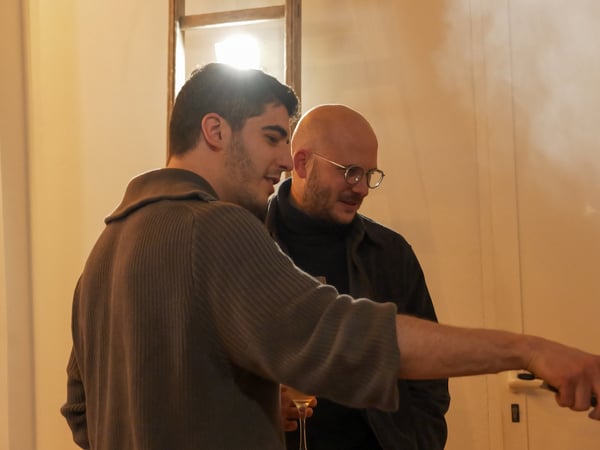 Two men are engaged in a social interaction indoors, with one man pointing towards something off-camera while the other looks on.