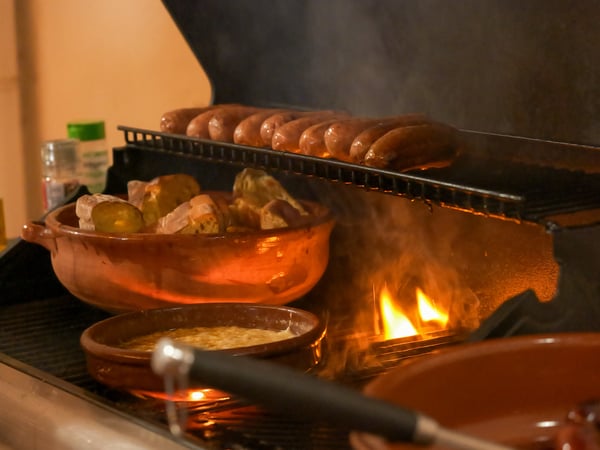 A photograph showing sausages grilling on a metal rack over an open flame, with bread and other dishes in rustic clay pots on the grill.