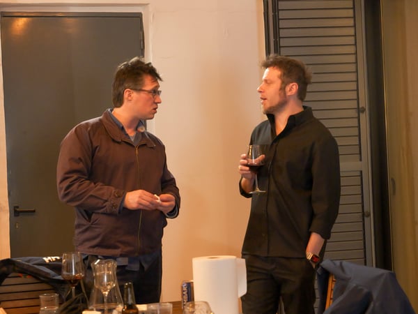 Two men are engaged in a conversation at an indoor social gathering, with one man holding a glass of red wine.