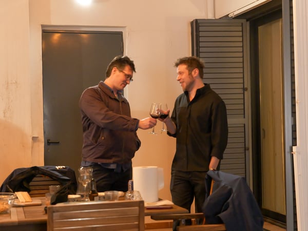 Two men are standing on a patio at night, smiling and clinking their red wine glasses together in a toast.